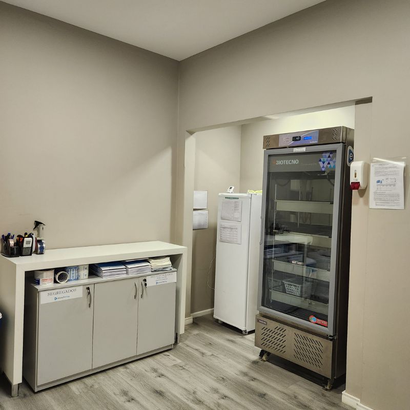 Sala com refrigerador vertical para medicamentos e armário identificado como refrigerados.